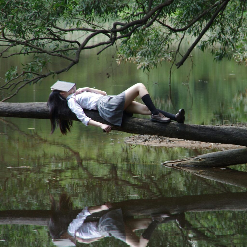 A young woman with a book on her face lies relaxing on a tree trunk by a tranquil lake.