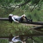 Micehelium G Vibes A young woman with a book on her face lies relaxing on a tree trunk by a tranquil lake.