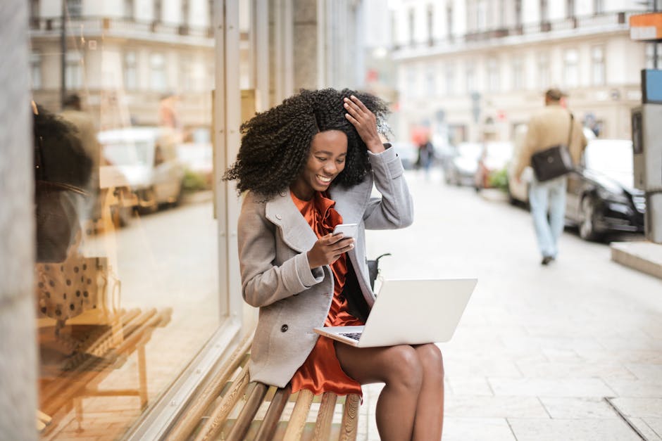 Smiling woman in an orange dress using laptop and smartphone outdoors, embracing remote work lifestyle.