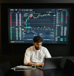 A focused man analyzing stock market trends on a laptop in an office setting with charts displayed.